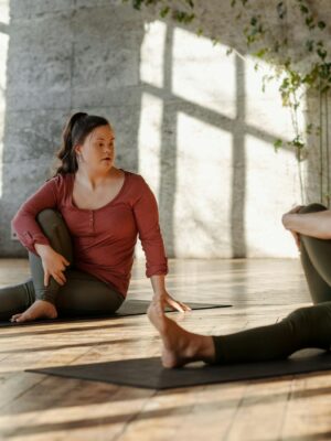 Two women practice yoga indoors on a sunny day, enhancing well-being and flexibility.